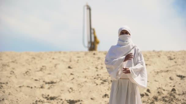 Араб в белой одежде, стоящий в пустыне, нефтяная вышка на заднем плане, топливный бизнес — стоковое видео