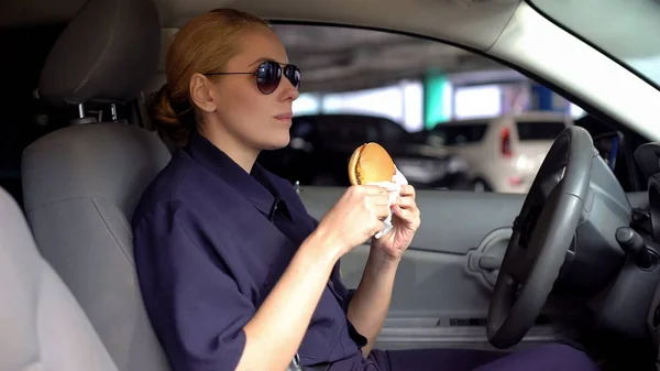 Female cop holding burger in police car, lack of time to healthy diet, junk food