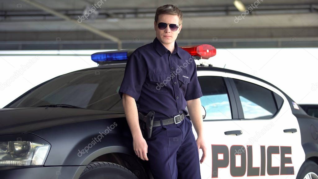 Serious policeman in sunglasses standing near patrol car, dangerous profession