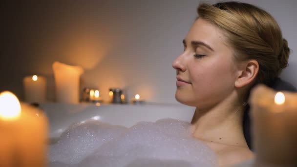 Jeune femme relaxante dans un bain chaud avec des bulles de mousse et des bougies, soirée — Video