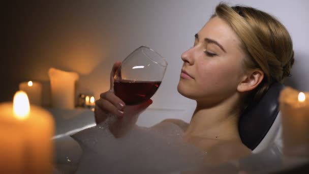 Ελκυστική γυναίκα που βρίσκεται στο μπάνιο με φυσαλίδες αφρού και κεριά, πίνοντας κρασί — Αρχείο Βίντεο