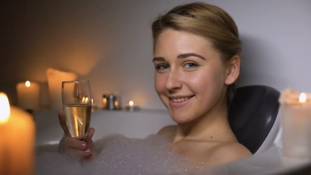 Fille dans le bain avec de la mousse et des bougies tenant un verre de champagne, souriant à la caméra — Video