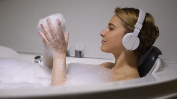 Mujer en auriculares jugando con burbujas de espuma en el baño, escuchando música de baile — Vídeos de Stock