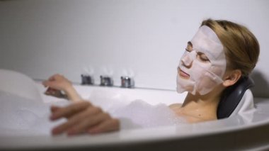 Köpük kabarcıkları ile banyoda rahatlatıcı genç kadın, yüzünde doğal esans maskesi