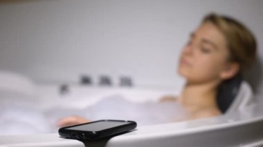 Köpük kabarcıkları ile banyoda yatan beceriksiz kadın, akıllı telefon suya düşen