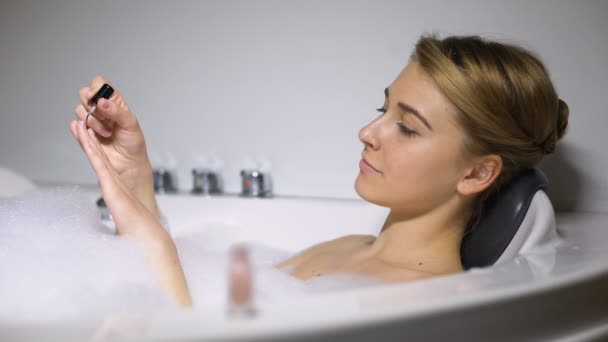 Mulher deitada no banho com bolhas de espuma, aplicando esmalte, procedimento de beleza — Vídeo de Stock