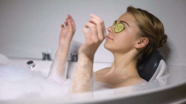 Κυρία δροσίζει στο μπάνιο με φυσαλίδες αφρού και αγγούρια στα μάτια, κλείνει το μάτι στην κάμερα — Αρχείο Βίντεο