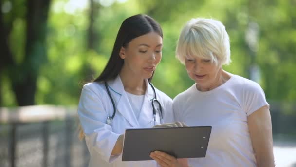 Lachende verpleegster toont ouderen vrouw examen resultaten in Hospital Park — Stockvideo