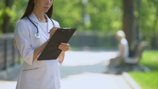 Operatore medico che compila dati medici nel parco, donna che fuma sullo sfondo — Video Stock