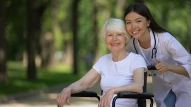 Tekerlekli sandalyedeki hemşire ve yaşlı kadın kameraya gülümsüyor ve başparmak-up gösteren