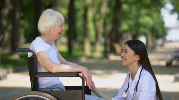 Infirmière souriante parlant avec une femme âgée handicapée dans le parc, centre de réadaptation — Video
