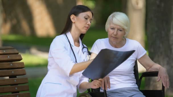 Doctora que muestra resultado de radiografía de columna a anciana discapacitada, día en el parque — Vídeo de stock