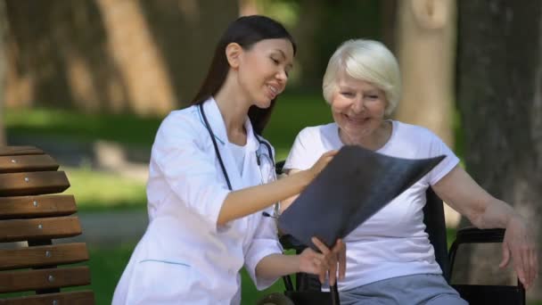 Sorridente medico femminile che mostra la radiografia spinale a felice donna anziana in sedia a rotelle — Video Stock