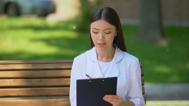 Sonriente doctora llenando datos en el banco en el parque hospitalario, trabajando al aire libre — Vídeo de stock