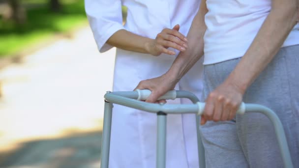 Женщина-медсестра поддерживает старушку-инвалида с прогулочной рамкой на открытом воздухе, реабилитационный центр — стоковое видео