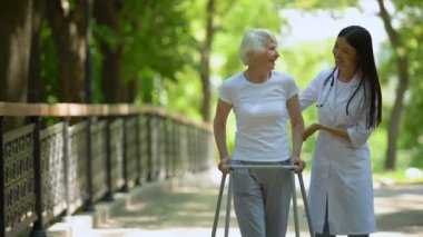 Kadın doktor yürüyüş çerçevesi ile yaşlı kadın yardım, hastane parkı, açık havada