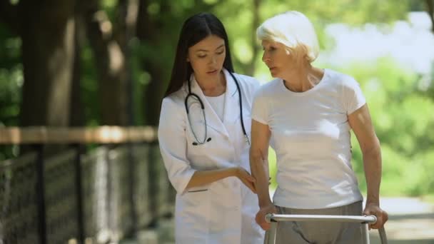 Медсестра поддерживает старушку, передвигающуюся по парку с помощью ходунков — стоковое видео