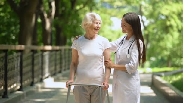 Médico femenino abrazando a una anciana con marco para caminar y sonriendo a la cámara al aire libre — Vídeo de stock