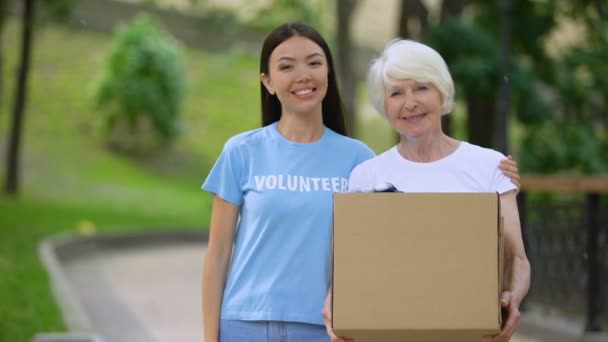 Заботливая активистка обнимает старушку за коробку для пожертвований, социальное волонтерство — стоковое видео