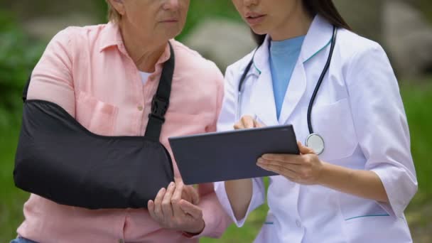 Terapeuta donna che mostra le immagini tablet alla vecchia signora ferita in fionda braccio, riabilitazione — Video Stock