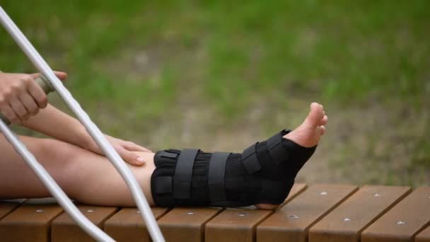 Femme avec béquille banc assis avec attelle de cheville sur la jambe, fracture osseuse, tension — Video