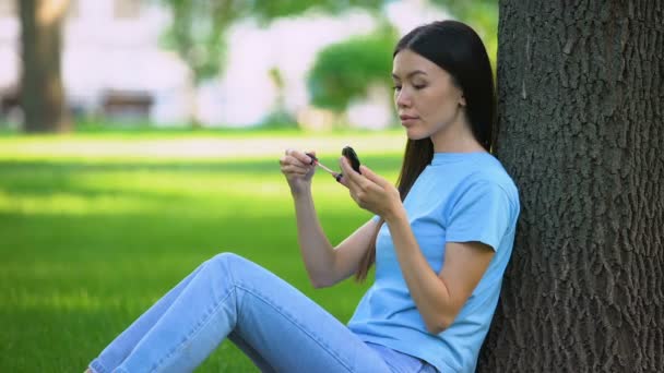 Vrij Aziatische vrouw toepassing lip gloss glimlachen kleine spiegel zittend Park, schoonheid — Stockvideo