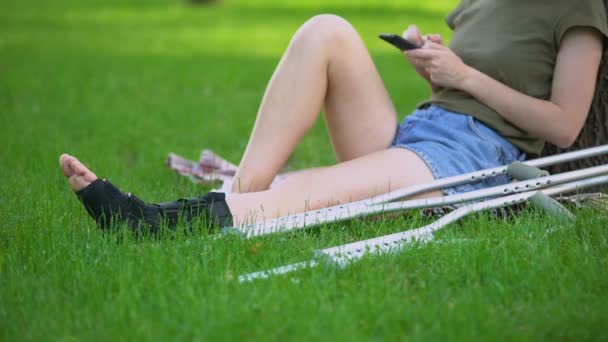 Mujer en tobillo sujetador sentado parque con muletas de desplazamiento smartphone, fractura — Vídeo de stock