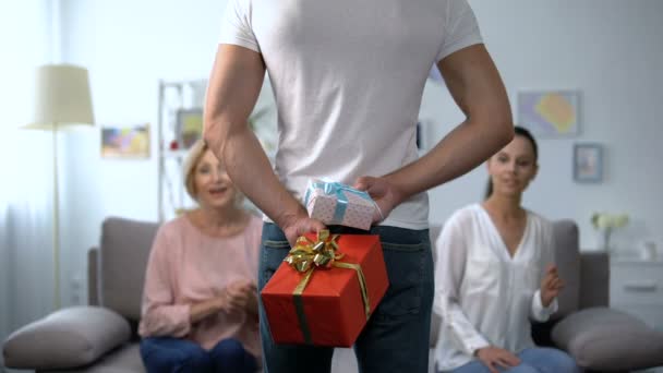 Curieux épouse et belle-mère regardant l'homme tenant des boîtes-cadeaux derrière le dos — Video