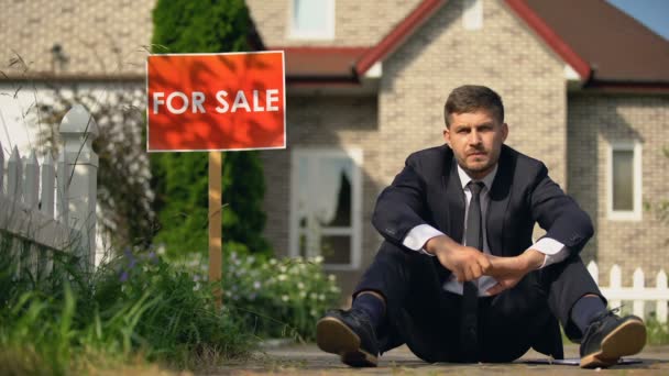 Депресивний чоловік, що сидить на землі біля знака продажу, втрата будинку через борги — стокове відео