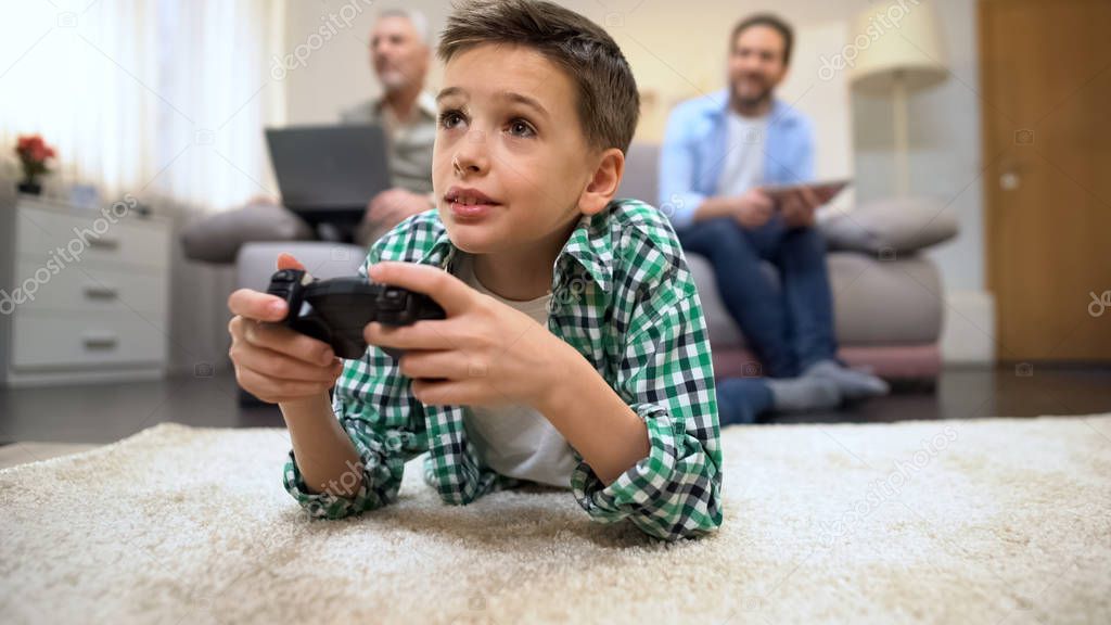 Little boy playing video game lying on floor, father and grandfather smiling
