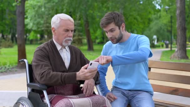 Молода людина пояснює пенсіонеру-інваліду, як користуватися смартфоном на відкритому повітрі — стокове відео