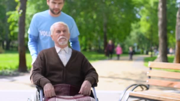 Мужчина-волонтер заботится о пожилом пациенте с ограниченными возможностями, проводит время в парке — стоковое видео