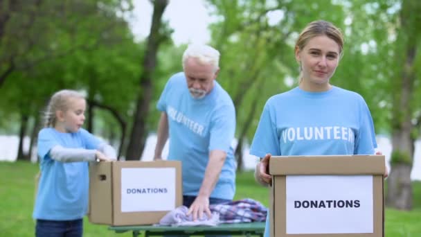 Pretty female volunteer holding box, activists sorting clothes on background — Stock Video