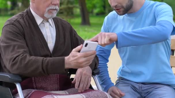 Sonriente voluntario masculino explicando anciano discapacitado cómo utilizar aplicación de teléfono, cuidado — Vídeo de stock