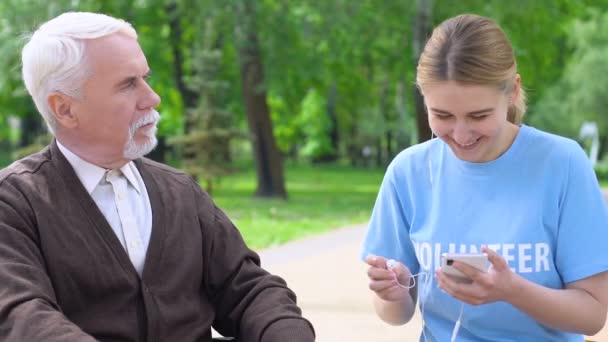 老いぼれた男にヘッドフォンを与える女性ボランティア、公園で音楽を聴く — ストック動画