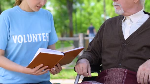 Uppriktig socialarbetare läsa bok till handikappade pensionär i Park, volontärarbete — Stockvideo