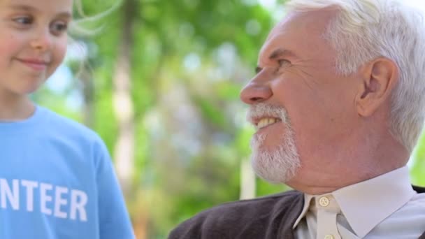 Маленькая девочка-волонтер разговаривает с одиноким пожилым мужчиной во время прогулки в парке, ухода за пенсионерами — стоковое видео