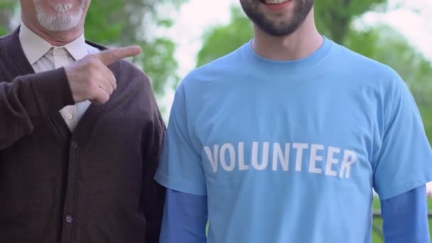 Літній чоловік і соціальний працівник вказують пальцем на футболку з логотипом волонтера — стокове відео