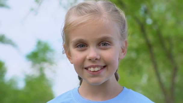 Χαριτωμένο θηλυκό παιδί ειλικρινά χαμογελά στην κάμερα, στέκεται στο πάρκο, χαρούμενη παιδική ηλικία — Αρχείο Βίντεο