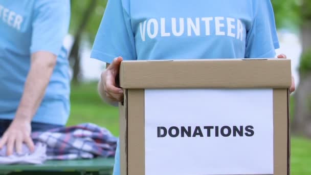 Mujer voluntaria sosteniendo caja de donaciones, personas clasificando ropa, ayuda humanitaria — Vídeo de stock