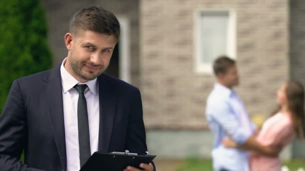 Agent immobilier professionnel caméra souriante, couple heureux étreinte arrière-plan — Video