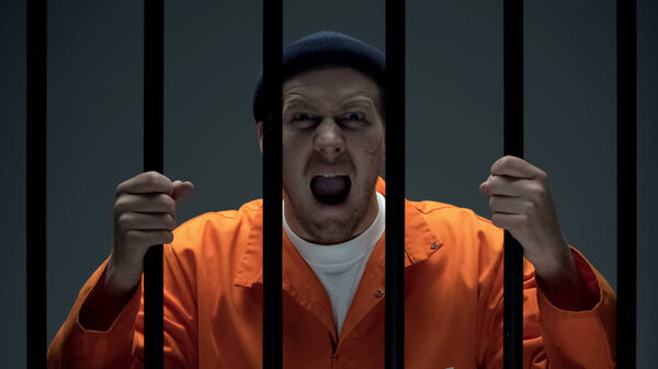 Агрессивный опасный заключенный мужчина со шрамом на лице, держащий решетки и кричащий
