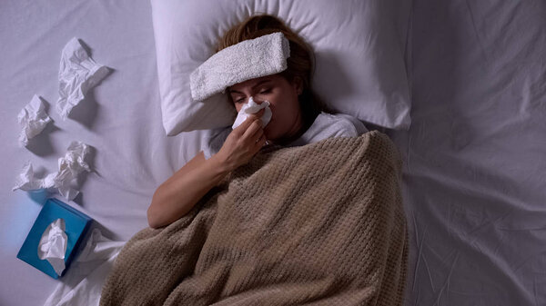 Sick woman with compress and napkins lying in bed, sneezing, seasonal allergy