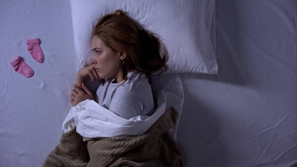 Грустная женщина плачет лежа в постели и глядя на детские носки возле подушки, выкидыш
