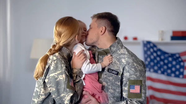 Cheerful military couple kissing little child in cheeks, USA flag on background