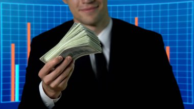 Successful man in suit showing bunch of dollars, business charts on background clipart