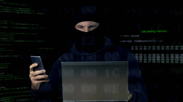 Verbrecher in Maske überprüft Überwachungskamera auf Laptop und Telefon, Datenbank — Stockbild Verbrecher Maske Überprüft Überwachungskamera Auf Laptop Und Telefon Datenbank — Stockfoto