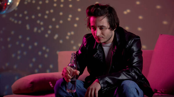 Pensive young man holding alcohol bottle at party, addiction problem, loneliness