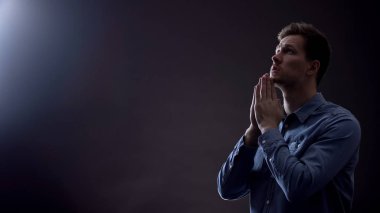 Young man praying in dark room under blessed light from heaven, Christianity clipart