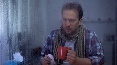 Mendil ile hasta adam yağmurlu pencere arkasında evde sıcak çay içme, grip